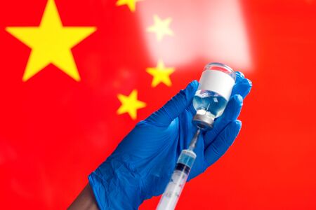 A doctor in blue medical gloves holding a bottle with vaccine and syringe in front of flag of China. Close up shot.の写真素材