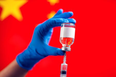 A doctor in blue medical gloves holding a bottle with vaccine and syringe in front of flag of China. Close up shot.の写真素材