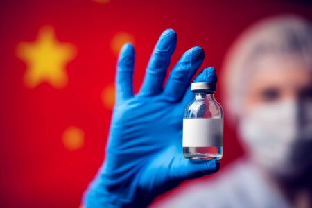 A doctor in blue medical gloves and mask holding in hand an ampula with vaccine in front of flag of China. Close up shot.の写真素材