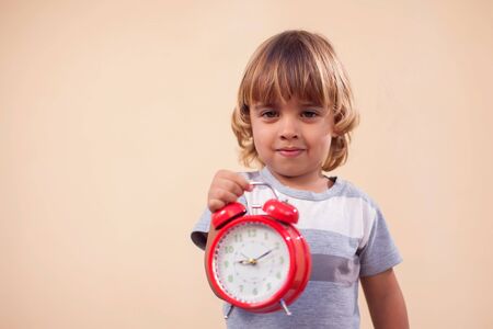 A portrait of kid boy holding red alarm clock. Childhood and time management concept