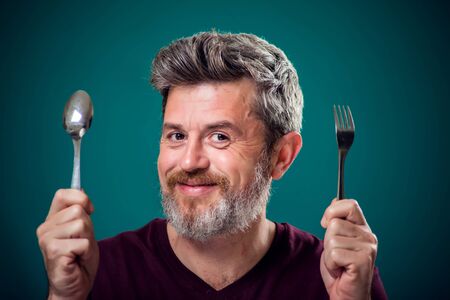 A portrait of bearded hungry man holding spoon and fork in hands. People and food concept