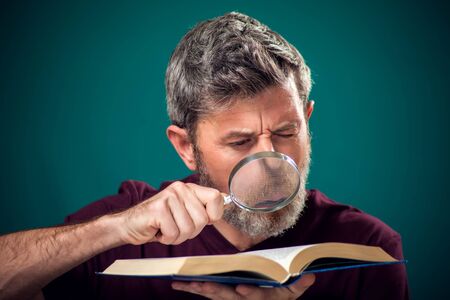 A portrait of bearded man in red t-shirt holding opened book and magnifying glass. People, education and searching conceptの写真素材
