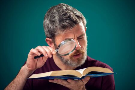 A portrait of bearded man in red t-shirt holding opened book and magnifying glass. People, education and searching conceptの写真素材