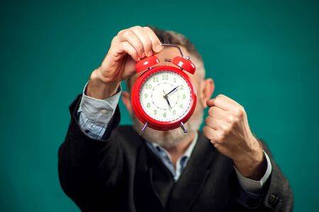 Angry bearded businessman in suit holding red alarm clock, shouting and showing fist at it. Time management conceptの写真素材