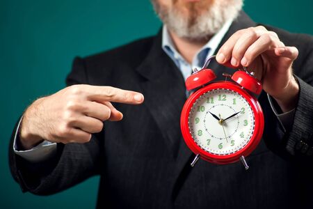 Bearded businessman in suit holding red alarm clock and pointing with a finger at it. Time management conceptの写真素材
