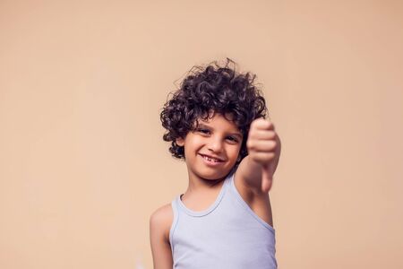 A portrait of smiling kid boy with curly hair showing thumb down gesture. Children and emotions conceptの写真素材
