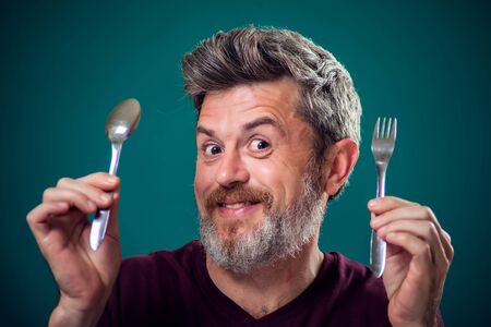 A portrait of bearded hungry man holding spoon and fork in hands. People and food concept