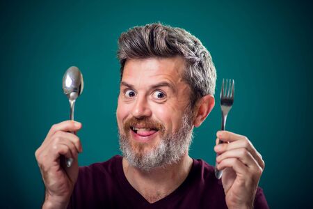 A portrait of bearded hungry man holding spoon and fork in hands. People and food concept
