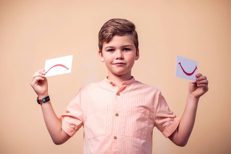 A portrait of kid boy holding positive and negative smile cards. Children and emotions conceptの写真素材