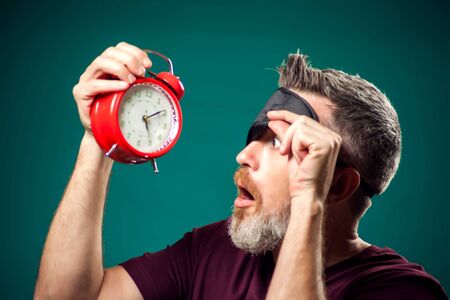 Surprised man in red t-shirt and sleep mask on head holding red alarm clock. Lifestyle and bed time conceptの写真素材