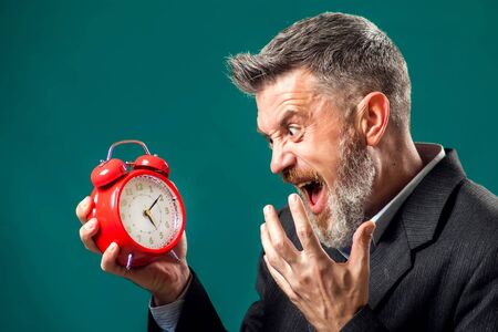 Angry bearded businessman in suit holding red alarm clock and shouting at it. Time management conceptの写真素材