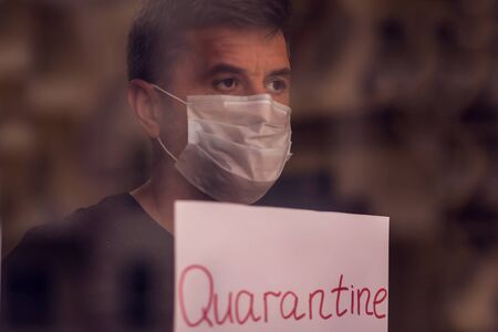A portrait of man indoor behind the window holding paper with word Quarantine. People, medicine and healthcare concept. Coronavirus epidemicの写真素材