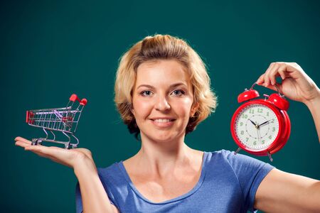 A portrait of smiling woman with short blond hair holding red alarm clock and shopping trolley. Shopping time and sales conceptの写真素材