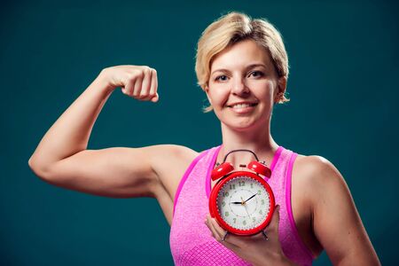A portrait of smiling woman with short blond hair in sportswear holding alarm clock. Fitness time...
