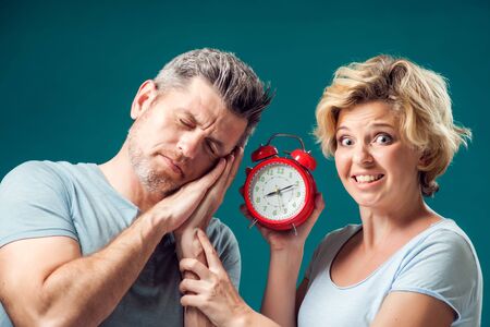 A portrait of couple in the morning. Woman holding alarm clock, while man sleeping, trying to wake...