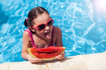 Kid girl showing tongue and holding watemelon in the swimming pool. Childhood, summer and holiday conceptの写真素材