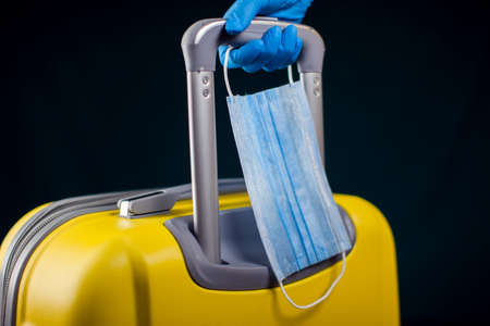 Luggage with medical face mask. Hand in medical gloves holding baggage. Travel and coronavirus concept. Studio shotの写真素材