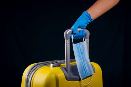 Luggage with medical face mask. Hand in medical gloves holding baggage. Travel and coronavirus concept. Studio shotの写真素材
