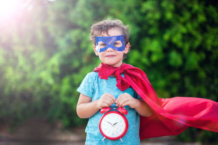 Superhero kid boy with alarm clock against green background outdoor. Childhood, success and and...