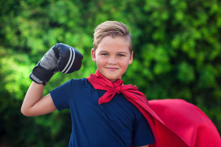 Kid boy superhero with boxing gloves and red cloak showing power outdoor. Childhood, success and and freedom conceptの写真素材