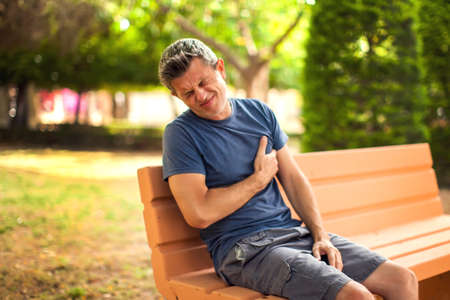 Man with heart pain outdoor. Male feeling chest pain sitting on the bench in the park. Healthcare and medicine conceptの写真素材