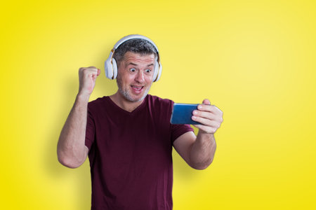 Man listening to the music or playing game on smartphone. Man with headphone holding cellphone on yellow backgroundの写真素材