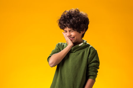 Portrait of kid boy having bad terrible toothache, holding hand on his cheek over yellow background....
