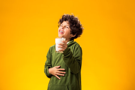 Child boy holding glass of milk and feeling abdominal pain over yellow background.の写真素材
