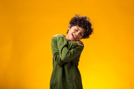 Portrait of smiling boy with closed eyes embracing himself on yellow backgroundの写真素材