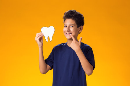 Portrait of child boy holding papercraft tooth and pointing finger at mouth, over yellow background....