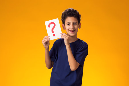 Portrait of thoughtful kid boy holding question mark card in hand over yellow background. Education...