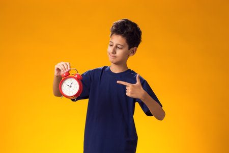 Portrait of child boy holding alarm clock and pointing with finger at it, over yellow background. Time management conceptの写真素材