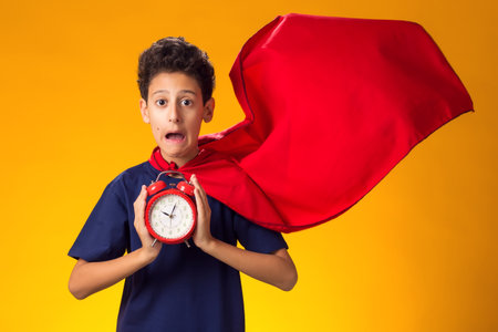 Portrait of surprised child plays superhero and holds alarm clock over yellow background. Education...