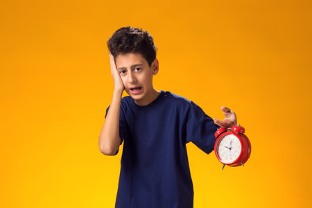 Portrait of surprised child boy holding alarm clock over yellow background. Time management conceptの写真素材