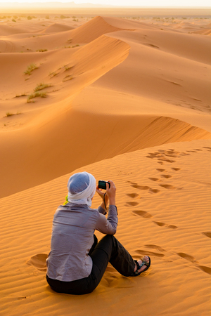 A woman is sitting on the sand dunes of Erg Chebbi and is photographing the Sahara sunrise. Merzouga, Moroccoの写真素材
