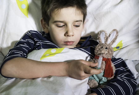 Sick child in bed with teddy bear. Measuring the temperature with a thermometer.