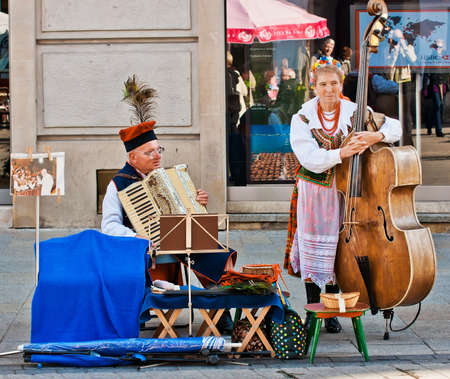 Street performers, an elderly woman and a man dressed in  polish folk costume playing traditional instruments, accordian and double bass, entertain tourists near the main city square in Krakow, Polandのeditorial素材