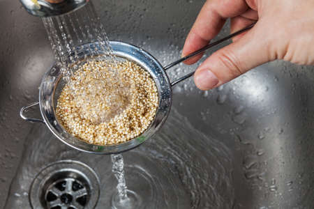 Man holding small collander with raw quinoa seeds and rinsing it in cold water in the kitchen sinkの写真素材