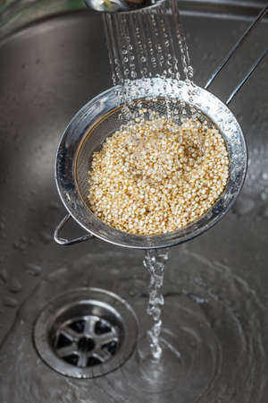 Man holding small collander with raw quinoa seeds and rinsing it in cold water in the kitchen sinkの写真素材