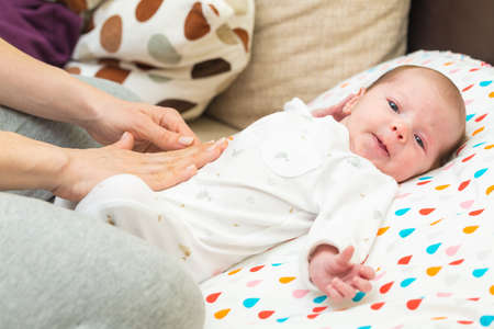 Newborn baby dressed in white laying on her back, with colics; mother's hand on her tummy
