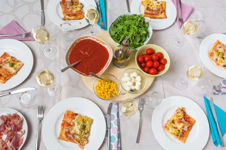 Top view of a dinner table with pizza slices on the plates and pizza ingredients in the middle: tomato sauce, arrugula, cherry tomato, mozzarella balls and corn; prosciutto on the side; plates with cutlery beside themの写真素材