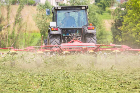 Farmer driving a tractor on the field with the attached tool used to spread hay on the field where it will dryの写真素材