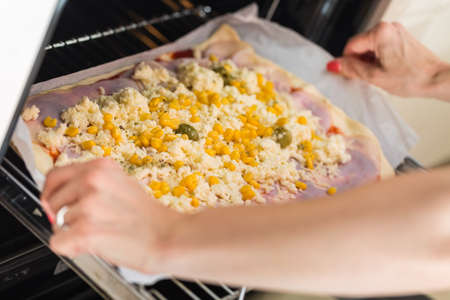 Woman's hands putting prepared homemade pizza with olive, ham, cheese and corn topping into the ovenの写真素材