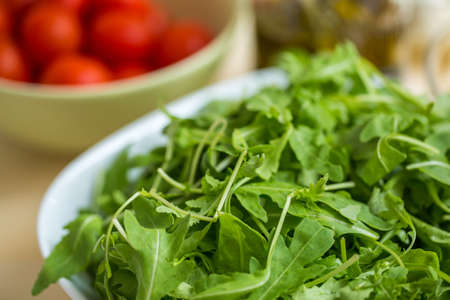 Fresh organic arrugula tossed in a white bowl, unseasoned, with cherry tomatoes in the backgroundの写真素材