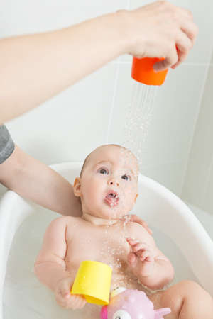 Seven months old baby girl in a bath, playing with pink toy and mothers arms around her, washing her and showering her from a plastic orange cupの写真素材