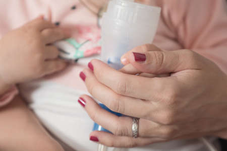 One year old baby girl inhaling from the inhaler, her mother holding her in the arms and comforting the ill babyの写真素材