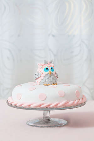 Birthday cake covered with fondant displayed on the pink cloth and glass tray; decorated with pink dots and an grey and pink fondant owl with blue eyes sitting on topの写真素材