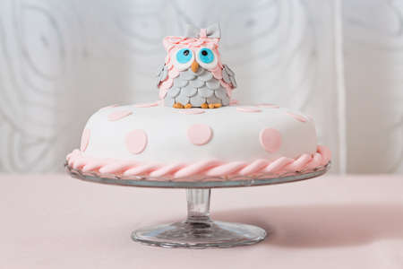 Birthday cake covered with fondant displayed on the pink cloth and glass tray; decorated with pink dots and an grey and pink fondant owl with blue eyes sitting on topの写真素材