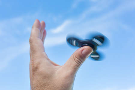 Man's hand holding a spinning fidget spinner in his hand, spinning them on his thumb, against the blue skyの写真素材