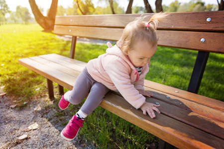 Eighteen months old toddler girl climbing on the wooden bench in the park on a sunny autumn dayの写真素材